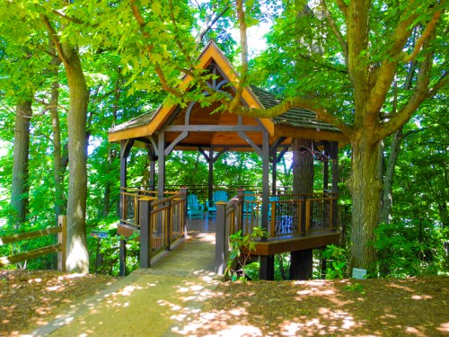 A wooden gazebo surrounded by lush green trees, featuring a pathway leading to it and blue chairs inside.