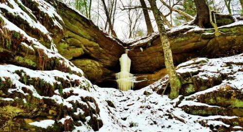 A frozen waterfall cascades down rocky cliffs, surrounded by snow-covered trees and a winter landscape.
