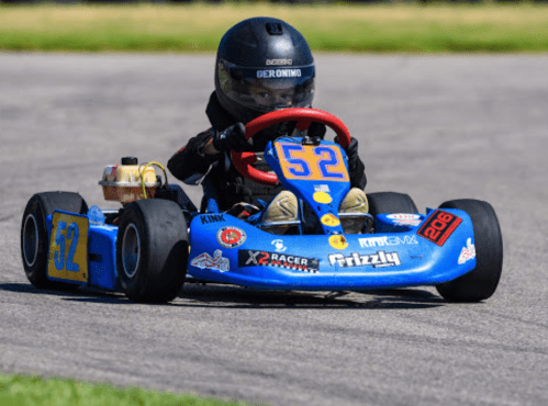 A young driver in a blue go-kart with the number 52 races on a track, wearing a black helmet and racing suit.