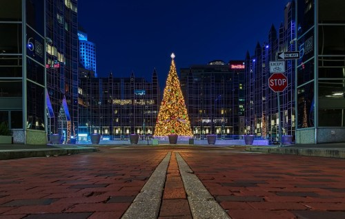 A brightly lit Christmas tree stands in a plaza at night, surrounded by modern buildings and festive decorations.
