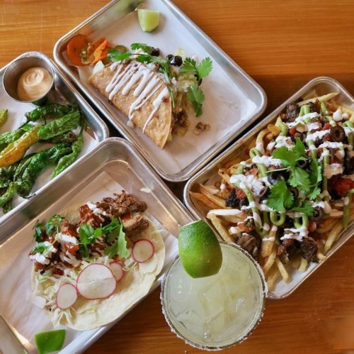 A vibrant spread of tacos, loaded fries, grilled peppers, and a margarita on a wooden table.