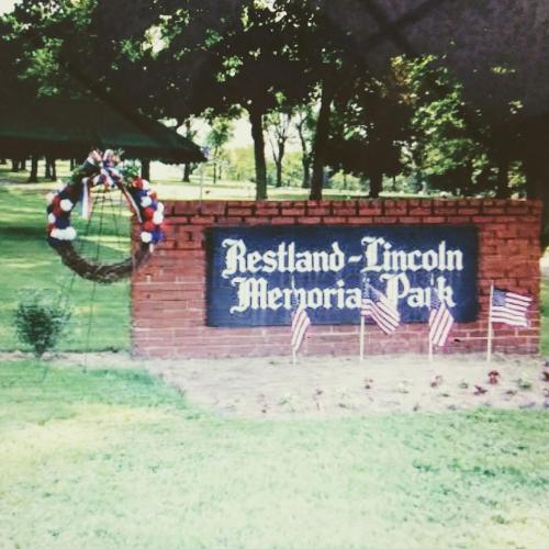 Sign for Restland-Lincoln Memorial Park, surrounded by greenery and small American flags, with a decorative wreath.