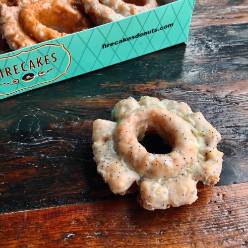 A glazed donut with a unique flower shape, resting on a wooden surface beside a teal box labeled "Firecakes."