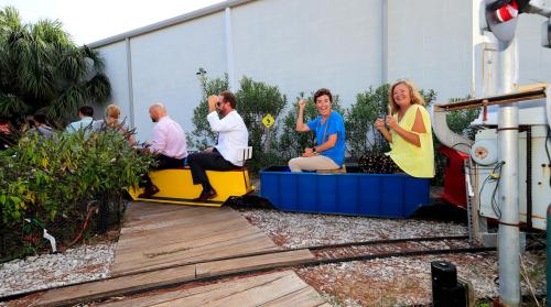 A small train with passengers smiles and waves, surrounded by greenery and a wooden pathway.