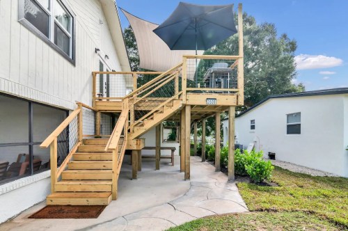 Wooden deck with stairs, shaded by an umbrella, next to a house and landscaped yard under a clear blue sky.