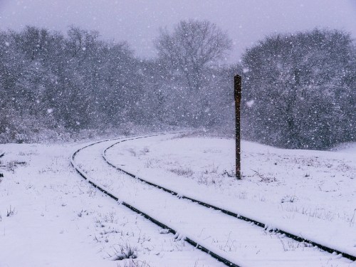 Snow-covered railway tracks curve through a winter landscape, surrounded by trees in a heavy snowfall.