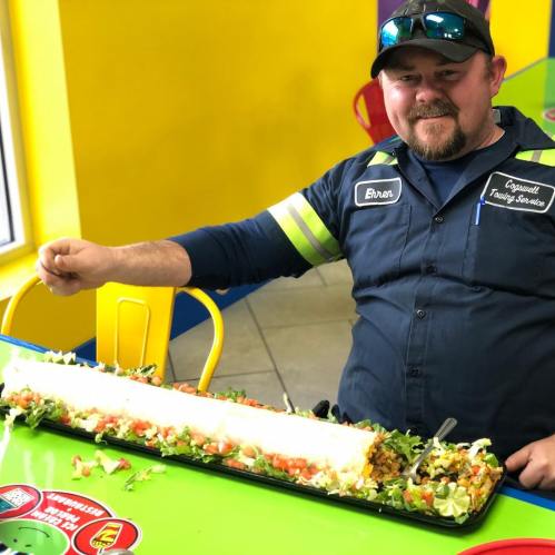 A man sits at a colorful table with a giant burrito filled with toppings, smiling proudly beside his meal.