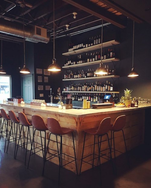A modern bar with a wooden counter, high stools, and shelves filled with wine bottles, illuminated by pendant lights.