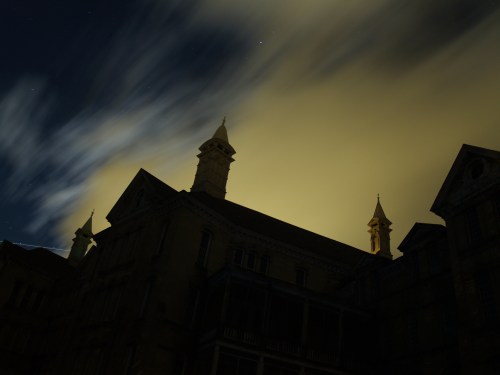 Silhouetted rooftops of a historic building against a cloudy night sky illuminated by a yellowish glow.