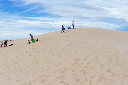 People enjoying a sandy dune under a blue sky with scattered clouds, some sitting and others climbing the hill.