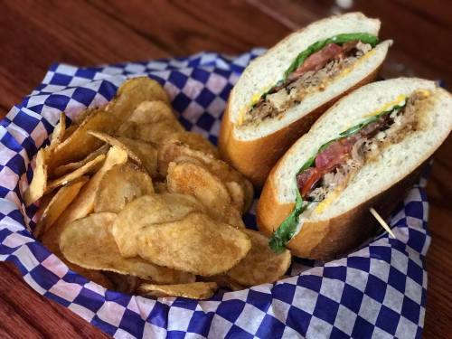 A basket with a sandwich filled with meat and greens, alongside a serving of crispy potato chips.
