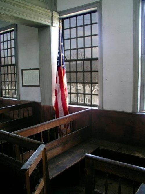 Interior of a historic room with wooden benches, an American flag, and large windows overlooking a graveyard.