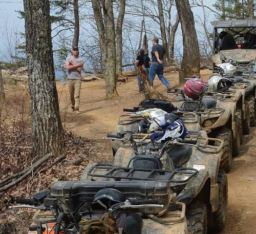 A line of parked ATVs on a dirt path in a wooded area, with people walking nearby.