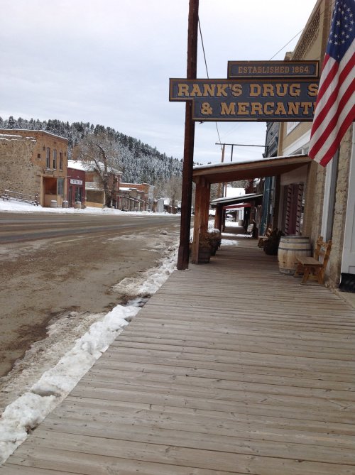 A snowy street scene featuring a wooden sidewalk and a sign for Rank's Drug Store & Mercantile in a historic town.