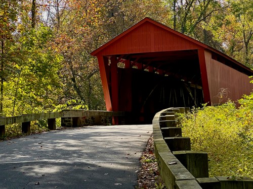A red covered bridge surrounded by autumn foliage and a winding road leading to it.