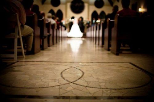 A wedding ceremony viewed from the back of a church, focusing on the couple at the altar and decorative floor tiles.