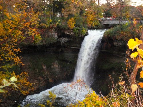 A waterfall cascades over rocky cliffs, surrounded by vibrant autumn foliage and trees.