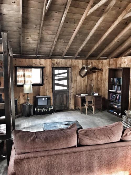 Cozy cabin interior with wooden walls, a sofa, a desk, a vintage TV, and a door leading outside.