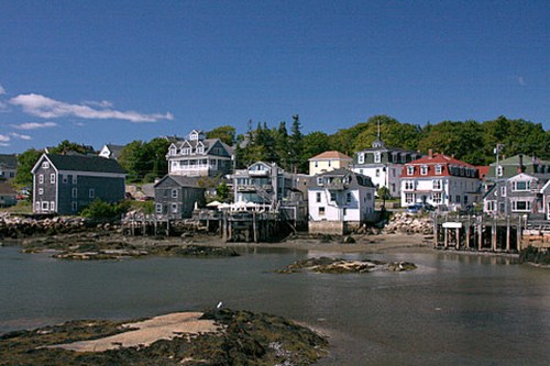 Coastal scene featuring colorful houses along the shore, with rocky outcrops and a clear blue sky.