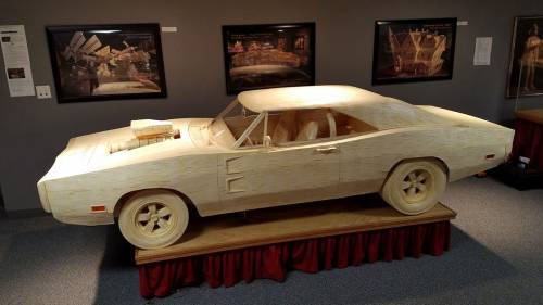 A wooden model of a classic car displayed on a pedestal, with art and information in the background.