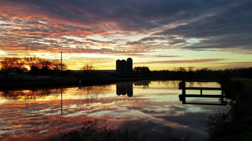 A serene sunset over a calm lake, reflecting vibrant colors and silhouettes of silos and trees.