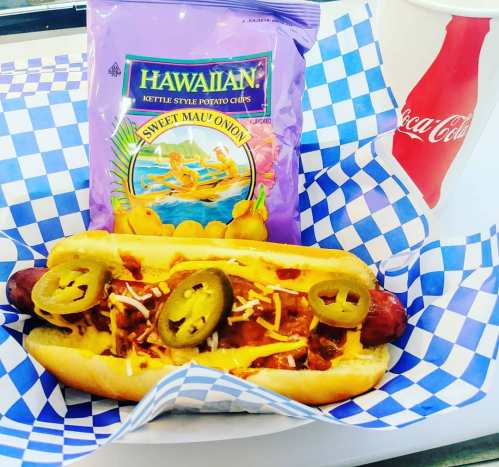 A hot dog topped with cheese, jalapeños, and chili, served with Hawaiian kettle chips and a soda in a checkered basket.
