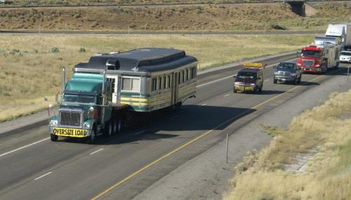 A large truck transports an oversized load, a vintage train car, on a highway with other vehicles nearby.