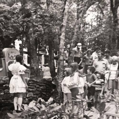A group of children and adults gather around a performer in a forested area, with trees and a stone structure in view.