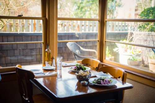 A cozy dining table set with plates and utensils, overlooking a sunny garden through large windows.
