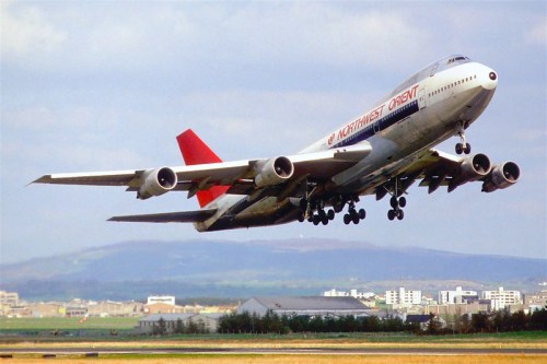 A Northwest Orient Airlines Boeing 747 takes off against a cloudy sky, with a cityscape in the background.
