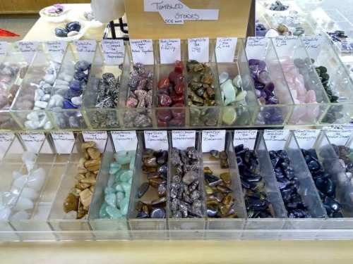 Display of various tumbled stones in clear containers, labeled with names, arranged by color and type.