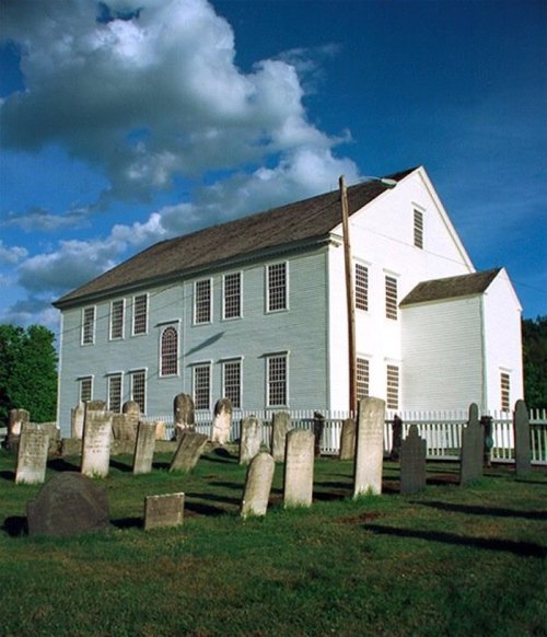 A historic white building beside a graveyard, with tombstones in the foreground and a blue sky with clouds above.