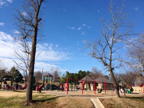 A playground with slides and swings, surrounded by trees and a clear blue sky, with children playing in the background.