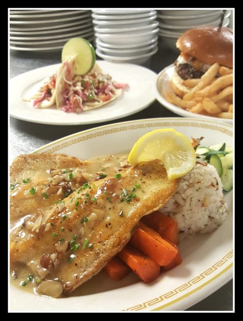 A plate of fish with lemon sauce, rice, and carrots, with tacos and a burger in the background.