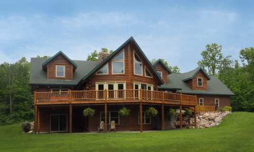 A large log cabin with a green roof, surrounded by trees and a grassy lawn, featuring a spacious deck.