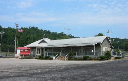 A sports facility with a gray building, flagpole, and surrounding fields under a clear blue sky.