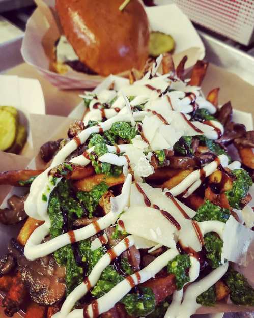 A plate of loaded fries topped with pesto, drizzled with sauce, and a burger in the background with pickles.