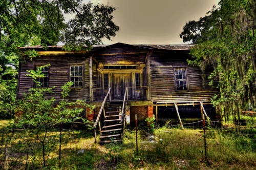 Abandoned wooden house with a front staircase, surrounded by overgrown grass and trees.