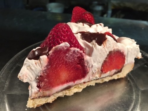 A slice of strawberry cream pie topped with fresh strawberries and chocolate drizzle on a clear plate.