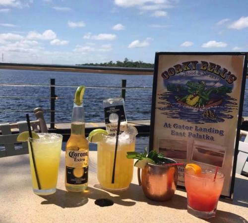 A table with four colorful cocktails and a menu, overlooking a scenic water view on a sunny day.