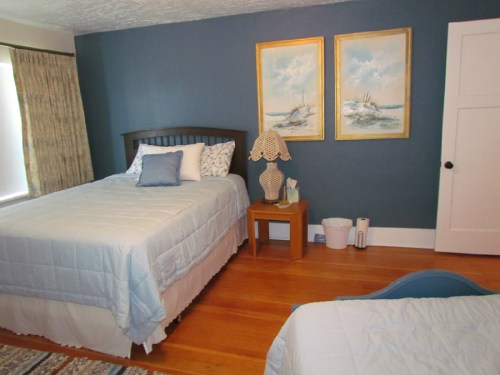 A cozy bedroom with two beds, blue walls, wooden floors, and framed paintings of ships.