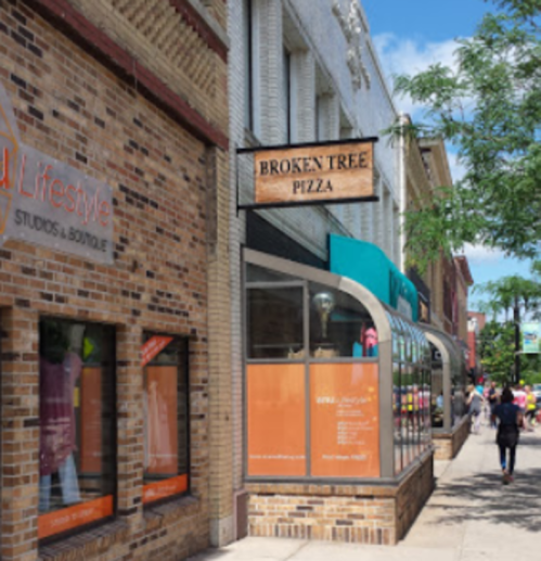 A street view of a pizza restaurant named "Broken Tree Pizza" next to a boutique, with people walking by.