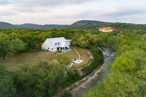 Aerial view of a modern white house surrounded by lush greenery and a winding river, with hills in the background.