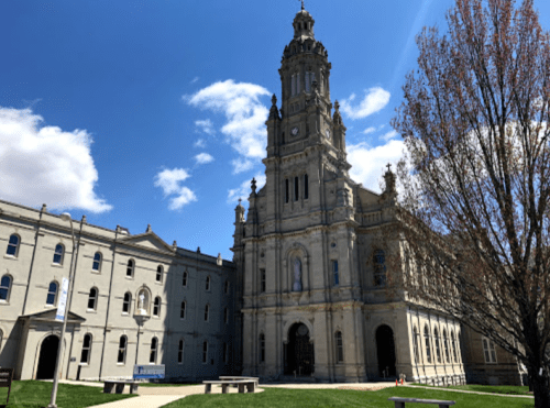 Historic building with a tall clock tower, surrounded by green grass and trees under a blue sky with clouds.
