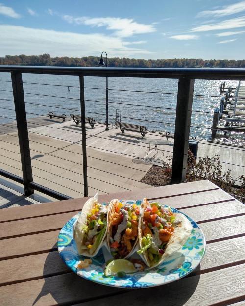 Three colorful tacos on a plate, with a scenic view of a river and dock in the background under a clear blue sky.