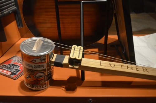 A homemade string instrument made from a can, wood, and strings, displayed in a showcase. The word "Luther" is visible.
