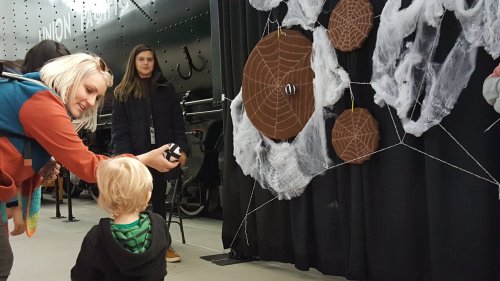 A woman interacts with a child in front of a spider web display, while another person stands in the background.