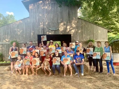 A group of people holding painted pumpkins in front of a rustic barn, with trees and grass in the background.