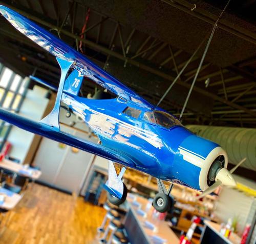A blue model airplane hanging from the ceiling in a restaurant, with wooden floors and tables visible below.