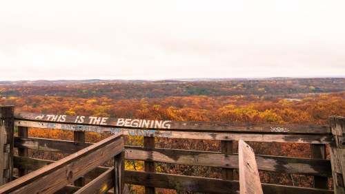 A wooden overlook with a view of colorful autumn trees under a cloudy sky, featuring graffiti that reads, "THIS IS THE BEGINNING."